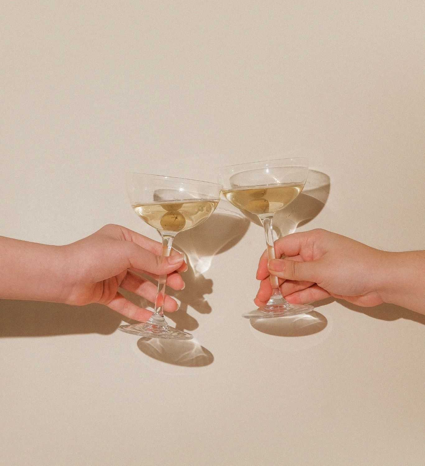 Two hands clinking cocktail glasses with olives against a beige background.