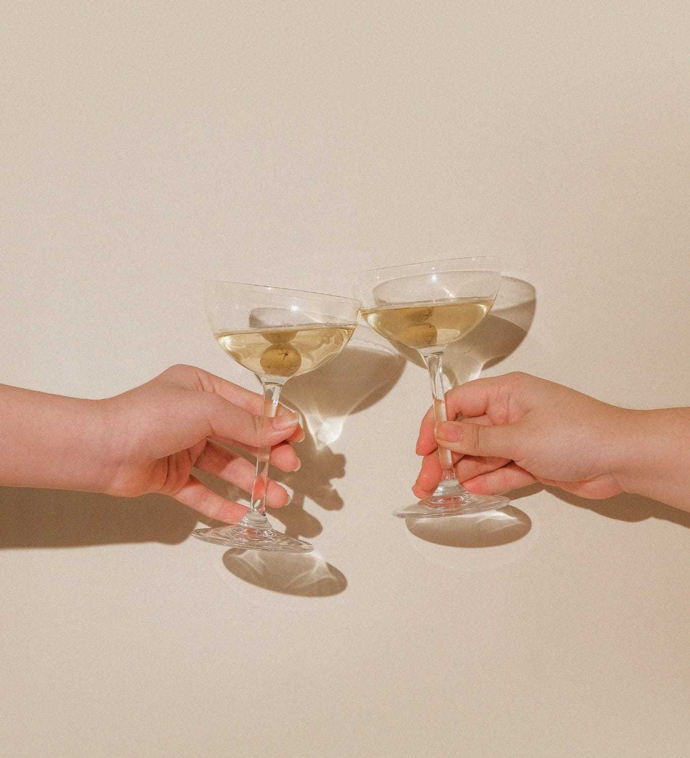 Two hands clinking cocktail glasses with olives against a beige background.